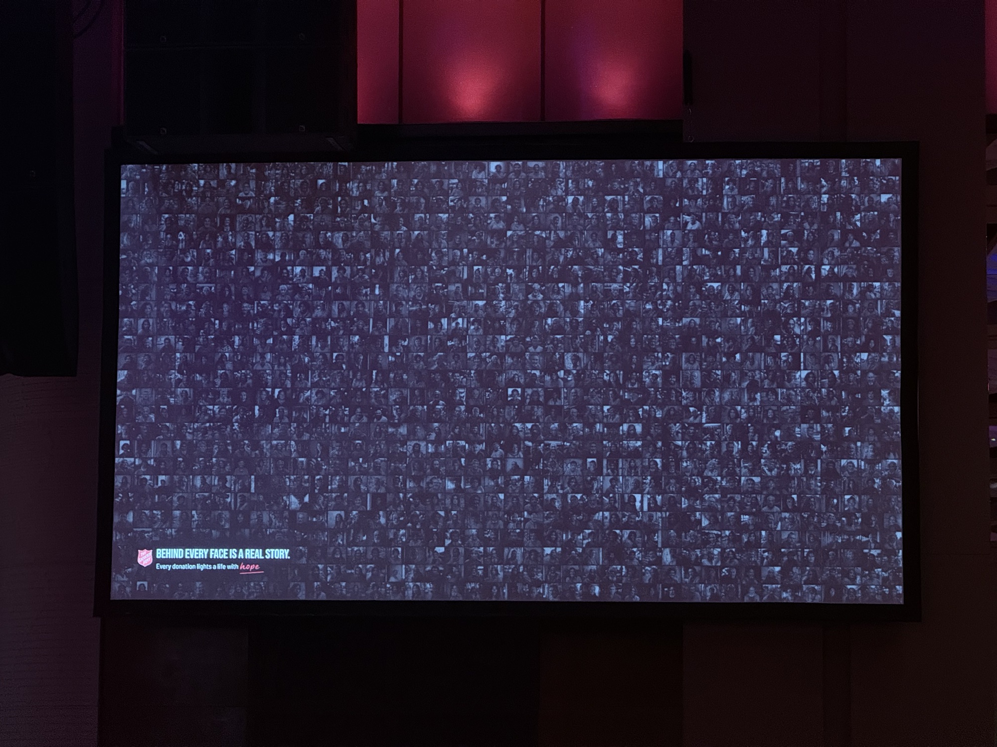 Close-up of the wall on a venue screen — hundreds of faces under 'Behind every face is a real story'